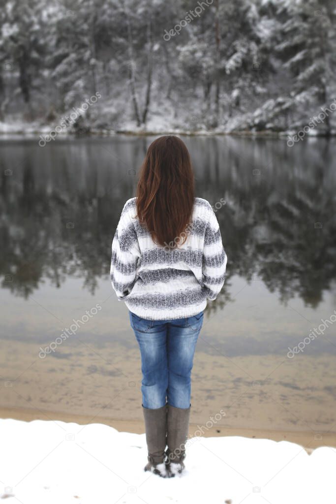 long-haired brunette in a gray sweater and blue jeans stands on the snowy shore of a winter forest river and looks at the trees reflected in the water