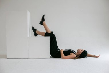 Siyah elbiseli, siyah ayakkabılı hoş, uzun saçlı bir kız beyaz bir stüdyoda yerde yatıyor.