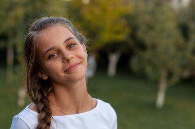 Uzun örgülü güzel bir kız yeşil bir bahçenin ortasında duruyor ve kameraya tatlı tatlı gülümsüyor.