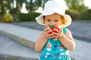 Komik bir gün mavi elbiseli bir kız çocuğu ve beyaz panama bir yaz parkında yolda duruyor ve oyuncak plastik domatesin tadına bakıyor.