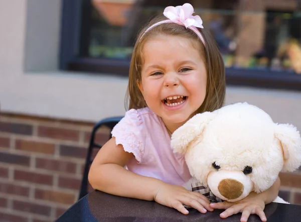 身穿粉红衣服头戴粉红花朵的黑头发婴儿坐在街边咖啡馆的桌子旁 抱着一只泰迪熊 开心地笑着 — 图库照片