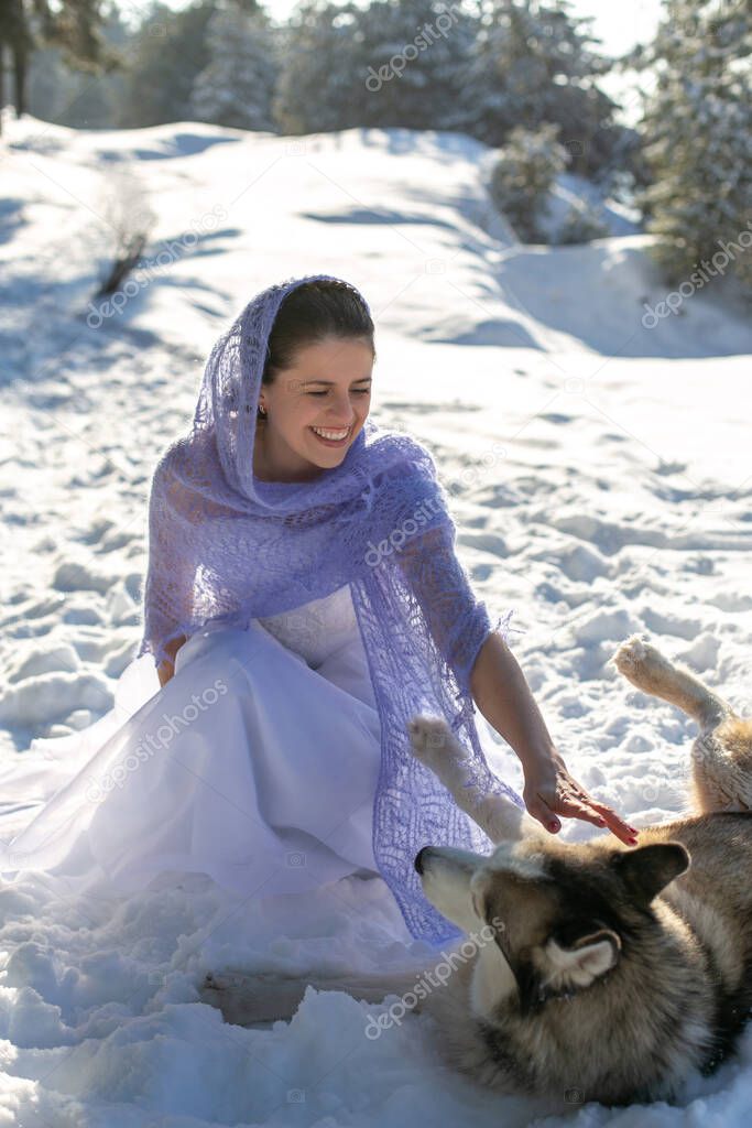 cute pretty cheerful girl in a white dress and a lilac shawl happily plays with a big gray dog in the winter forest