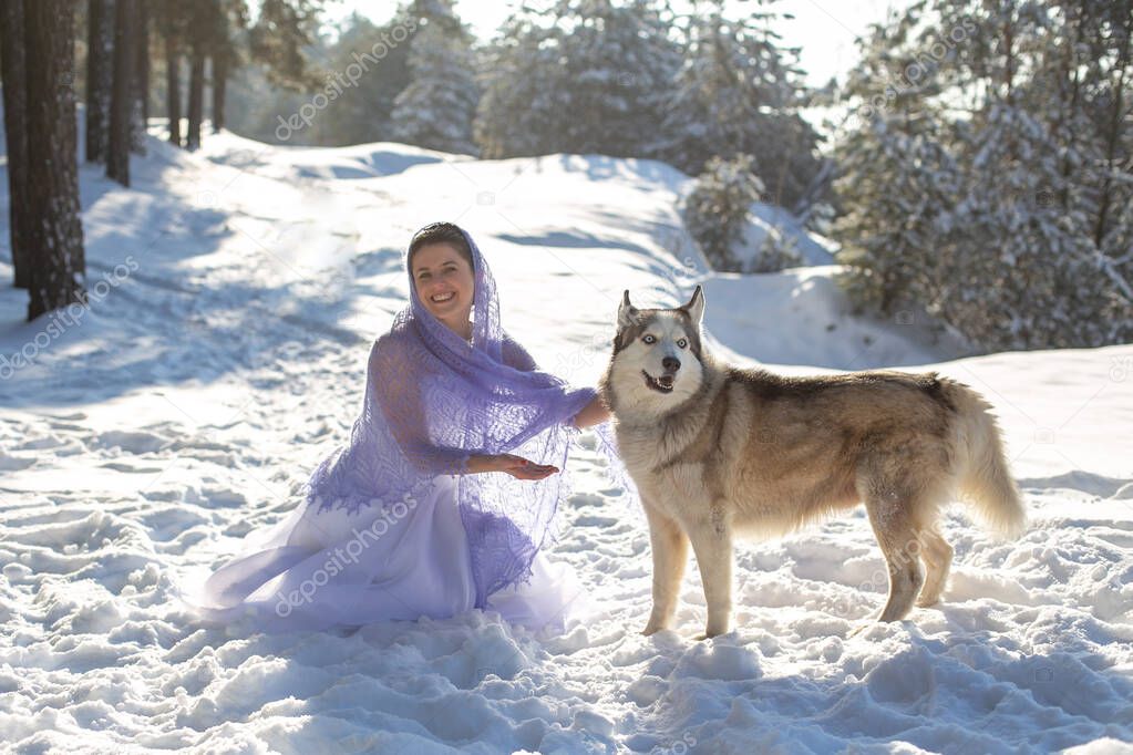 cute pretty cheerful girl in a white dress and a purple shawl happily stroking a large gray dog in the winter forest