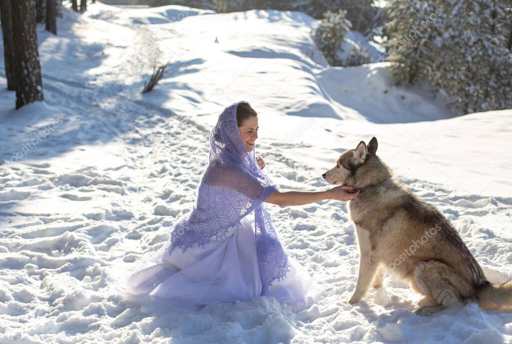 cute cheerful dark-haired girl in a long white dress and a light lavender openwork shawl happily stroking a large gray dog in the winter forest