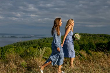 Mavi pamuklu elbiseli iki güzel kadın güzel bir gökyüzüne karşı otla kaplı uzun bir nehir kıyısında el ele yürüyorlar.