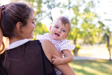 Küçük bebek annesinin kollarında oturuyor ve ilgiyle kameraya bakıyor.