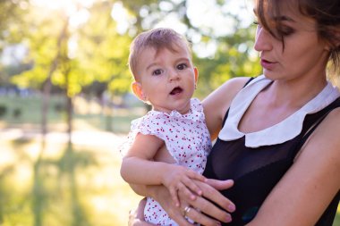 Küçük bebek annesinin kollarında oturuyor ve ilgiyle kameraya bakıyor.