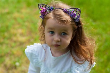 Kıvırcık saçlı şirin kız yaz parkındaki çimlerin üzerinde duruyor ve doğrudan kameraya bakıyor.