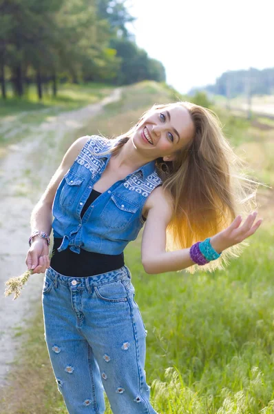 穿着牛仔裤衣服的金发美女站在一片夏日森林的郊外小径上 她的头发在风中飘扬 对着摄像机笑着 — 图库照片