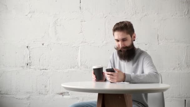 Glad skäggig man sitter vid ett skrivbord med telefon vid bordet och dricker kaffe — Stockvideo