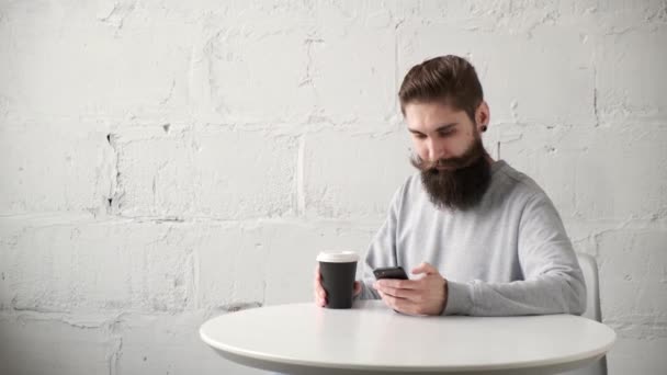 Szczęśliwy brodaty mężczyzna siedzi przy biurku z telefonem przy stole i pije kawę. — Wideo stockowe