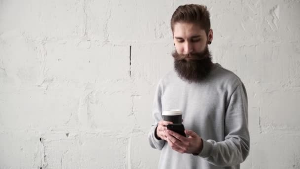 若い男がコーヒーを飲んで携帯電話を見て。幸せな髭の男が微笑む — ストック動画