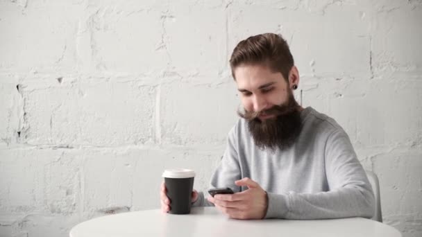 Glad skäggig man sitter vid ett skrivbord med telefon vid bordet och dricker kaffe — Stockvideo