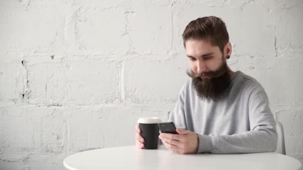 Glad skäggig man sitter vid ett skrivbord med telefon vid bordet och dricker kaffe — Stockvideo