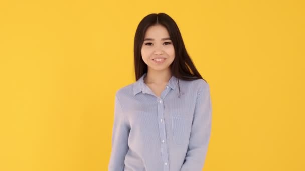 Una chica joven en una camisa azul sobre un fondo amarillo sonriendo y mostrando signo OK — Vídeos de Stock