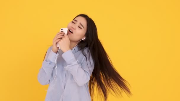 Азиатка в наушниках на желтом фоне поет караоке по телефону и танцует — стоковое видео