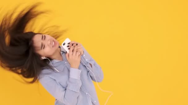 Азиатка в наушниках на желтом фоне поет караоке по телефону и танцует — стоковое видео