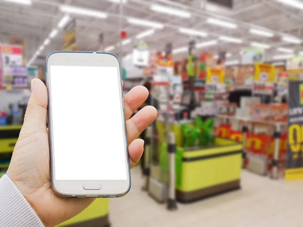 Left asian hand with pullover holding blank screen smart phone on blurred background of cash desk in supermarket — Stock Photo, Image