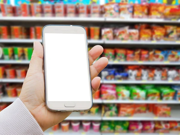 left asian hand with pullover holding blank screen smart phone on blurred background of various brand of instant noodles in packaging for sale on supermarket stand or shelf