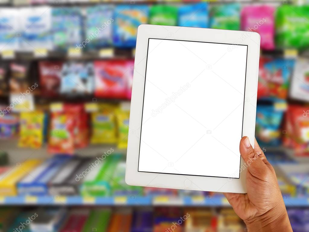 asian right hand holding blank tablet screen on blurred various brand of candy in packaging for sale on supermarket stand or shelf 