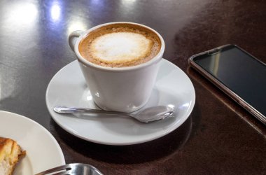 Bir fincan cappuccino, bir kafede latte, bir masanın üzerinde, siyah ahşap bir yüzey, akıllı telefonun yanında. Randevu, bekleme konsepti. Konsept kafede kahvaltı. Yatay yönelim.