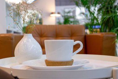 Yeşil bitkiler ve deri koltuklar olan açık bir kafedeki küçük bir masada bir fincan kahve ya da çay. Masada beyaz çiçekli beyaz vazo. Arka plan bulanık. Seçici odaklanma. Yatay yönelim