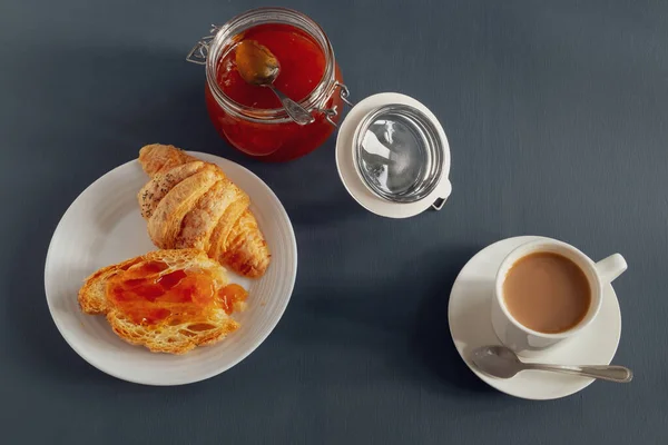 A jar of jam, a fresh croissant smeared with jam, and a cup of coffee with cream for breakfast or coffee pause on a dark gray background. Horizontal orientation. Selective focus. View from above.