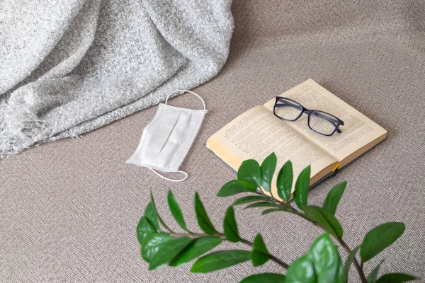 Otevřená kniha, brýle a lékařská maska leží na pohovce s kostkovaným — Stock fotografie