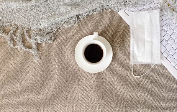 A cup of coffee, a keyboard, plaid and a medical mask are on the sofa. Self-isolation, quarantine, stay home concept. Horizontal orientation. Selective focus. View from above.