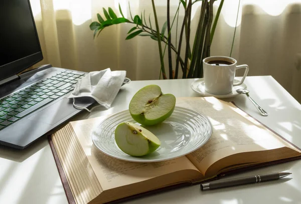 A workplace with a computer, an open book, an apple, a medical mask and a cup of coffee. Home office, remote work, distance education concept. Horizontal orientation, selective focus.