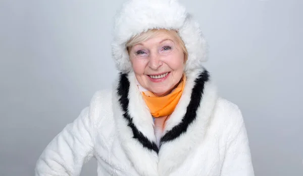 成人微笑的妇女是70岁 一个老妇人在轻的毛皮帽子和毛皮外套反对轻的背景 — 图库照片