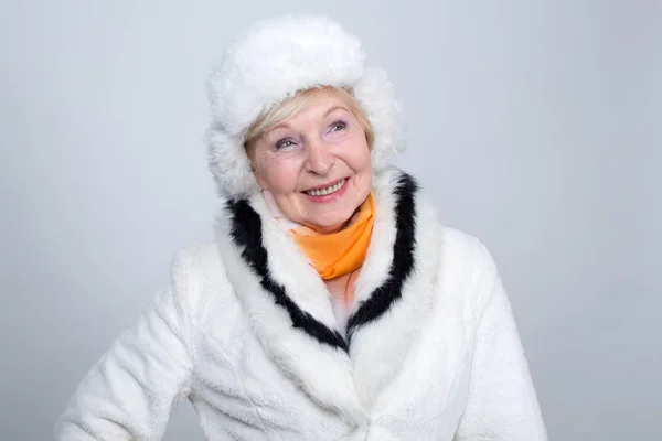 成人微笑的妇女是70岁 一个老妇人在轻的毛皮帽子和毛皮外套反对轻的背景 — 图库照片