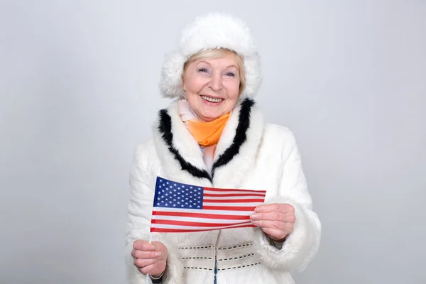 持有美利坚合众国国旗的高级妇女 成年妇女70岁 老女人穿着暖和的冬衣站在浅色的背景 — 图库照片