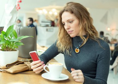 Kadın oturan bir kafe bir pencerenin önünde bir akıllı telefon elinde tutar. O kahve içer ve yazar veya iletileri veya bir sosyal ağ internet haber okur.