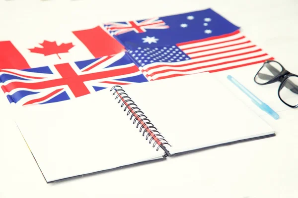 Education concept. English language learning. English speaking countries. Notebook, textbook, blue pen, glasses, four flags on a wooden table.