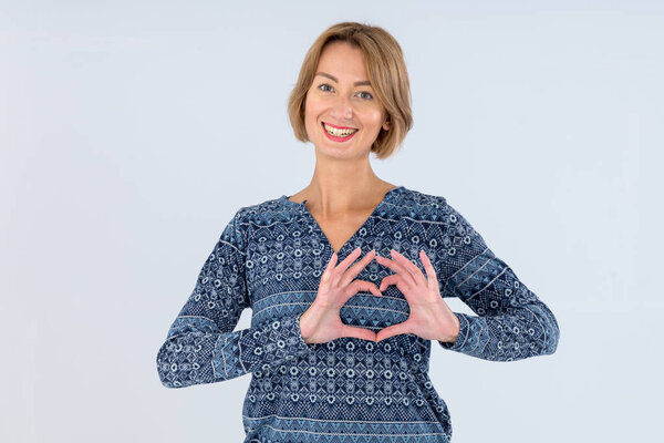 Молодая женщина показывает символ сердца своими руками, стоя на светло-сером фоне
.