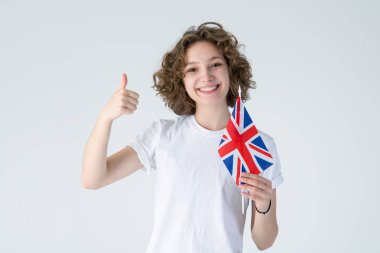 Hoşuma gitti! Uk bayrağıyla beyaz arka planda duran kıvırcık, genç bir kadın baş parmağını gösteriyor. İngilizce öğrenmek iyidir..