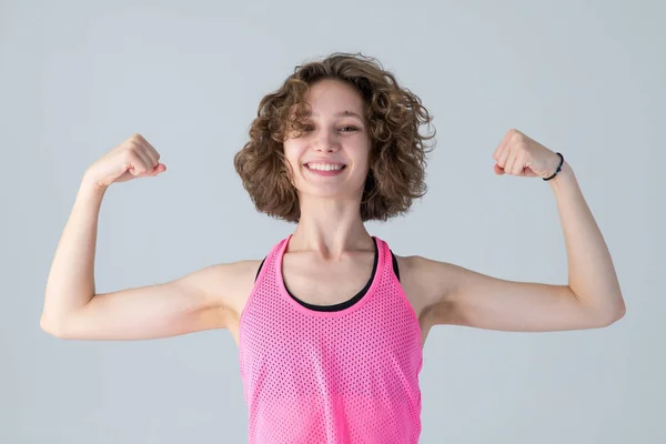 我很坚强 上健身课穿粉红衬衫的快乐女孩 卷曲的年轻女子举起双手 露出肌肉 站在灰色的背景上 — 图库照片