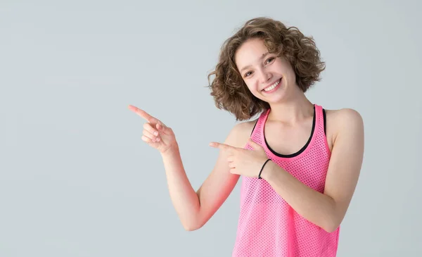 若い運動選手の女性は 灰色の背景に立って 側面に指をポイントします 幸せな女の子でAピンクシャツのためにフィットネスクラス — ストック写真