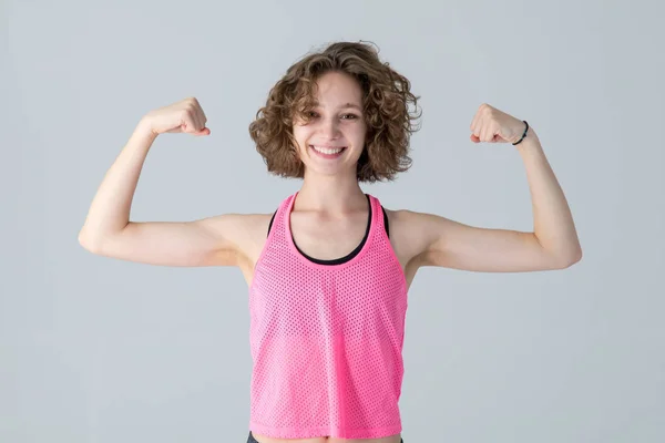 卷曲的年轻女子举起双手 露出肌肉 站在灰色的背景上 上健身课穿粉色衬衫的快乐女孩 — 图库照片