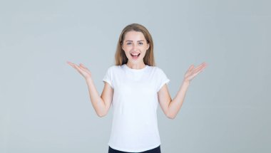 Vay canına! Gri arka planda tek başına duran komik çığlık atan genç kadın. Sıradan beyaz tişörtlü, sürpriz yüzlü ve kameralı bir kadın..