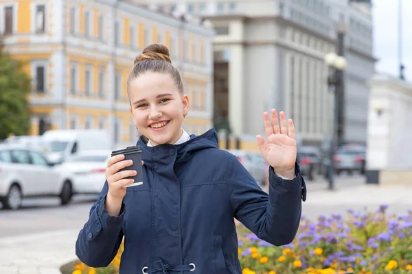 Good afternoon! Teenage girl shows a hello gesture or says goodbye and holds a cup of coffee in her hand.