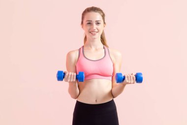 Elinde iki dambıl olan, pembe arka planda duran genç bir kadın. Fitness egzersizi.