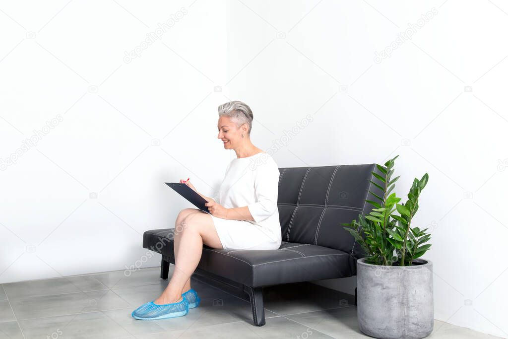 Mature woman is filling out a questionnaire while sitting on a sofa in a clinic reception room.