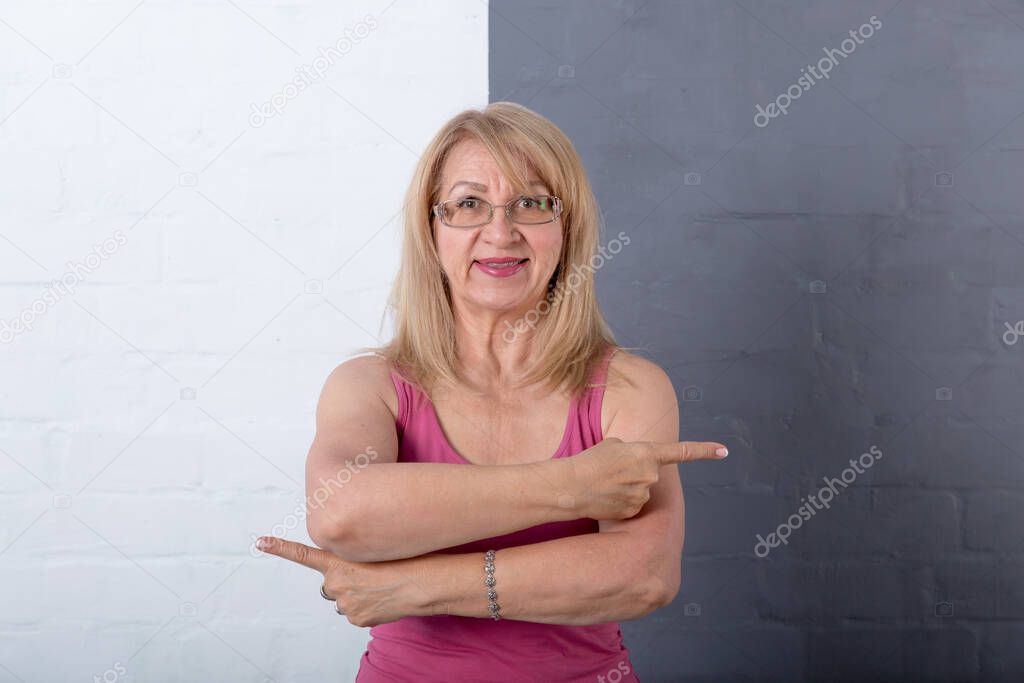 Happy mature woman points her fingers in different directions while standing against gray and white wall. Place for text comparison, fitness and diet.