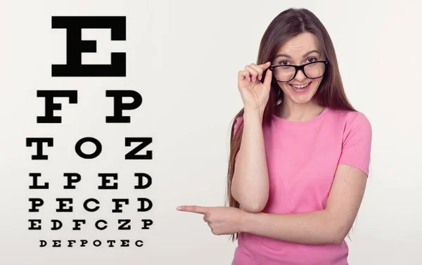 ビジョンの問題だ 眼鏡をかけた若い女性が目の検査のための文字のテーブルを指して カメラを見ています 眼科学 — ストック写真