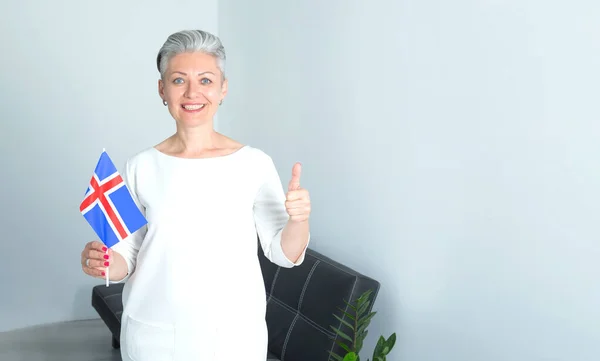 站在家里的房间里 拿着冰岛国旗的快乐而成熟的女人感激地举起了她的大拇指 谢谢你 一切都会好的 — 图库照片