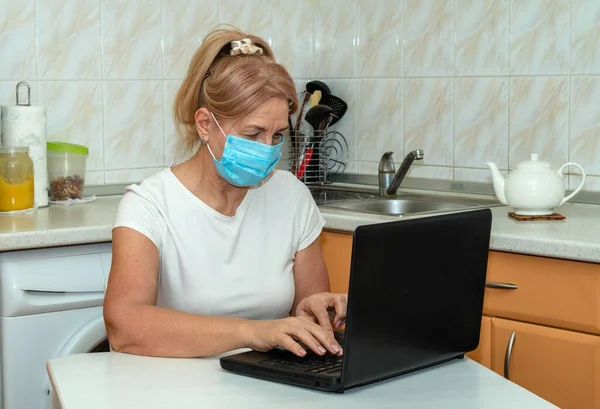 Maskeli yetişkin bir kadın mutfakta oturmuş dizüstü bilgisayarına bir mesaj yazıyor. Karantinada online iletişim - kavram. İnternet üzerinden bir doktora danışıyorum..