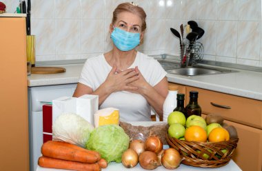 Tıbbi koruma maskesi takmış yetişkin bir kadın, ev yemeklerinin arka planında oturan yiyecek ve ürünlerin teslimatı için bir minnettarlık işareti gösteriyor. Karantina - evde kal.