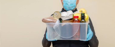 Koruyucu tıbbi maskeli ve eldivenli bir kurye plastik kutuda ürünleri teslim ediyor. Bağış kuruluşunda gönüllü çalışıyor. Koronavirüs karantinası sırasında yiyecek bağışı.